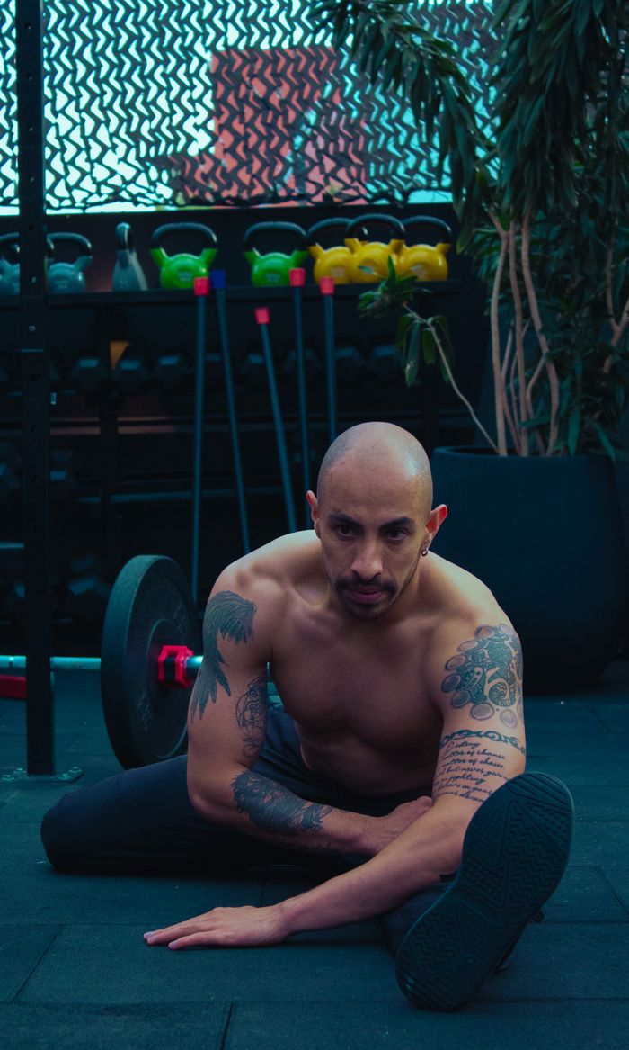 Shirtless tattooed man stretching in urban gym, surrounded by fitness equipment.