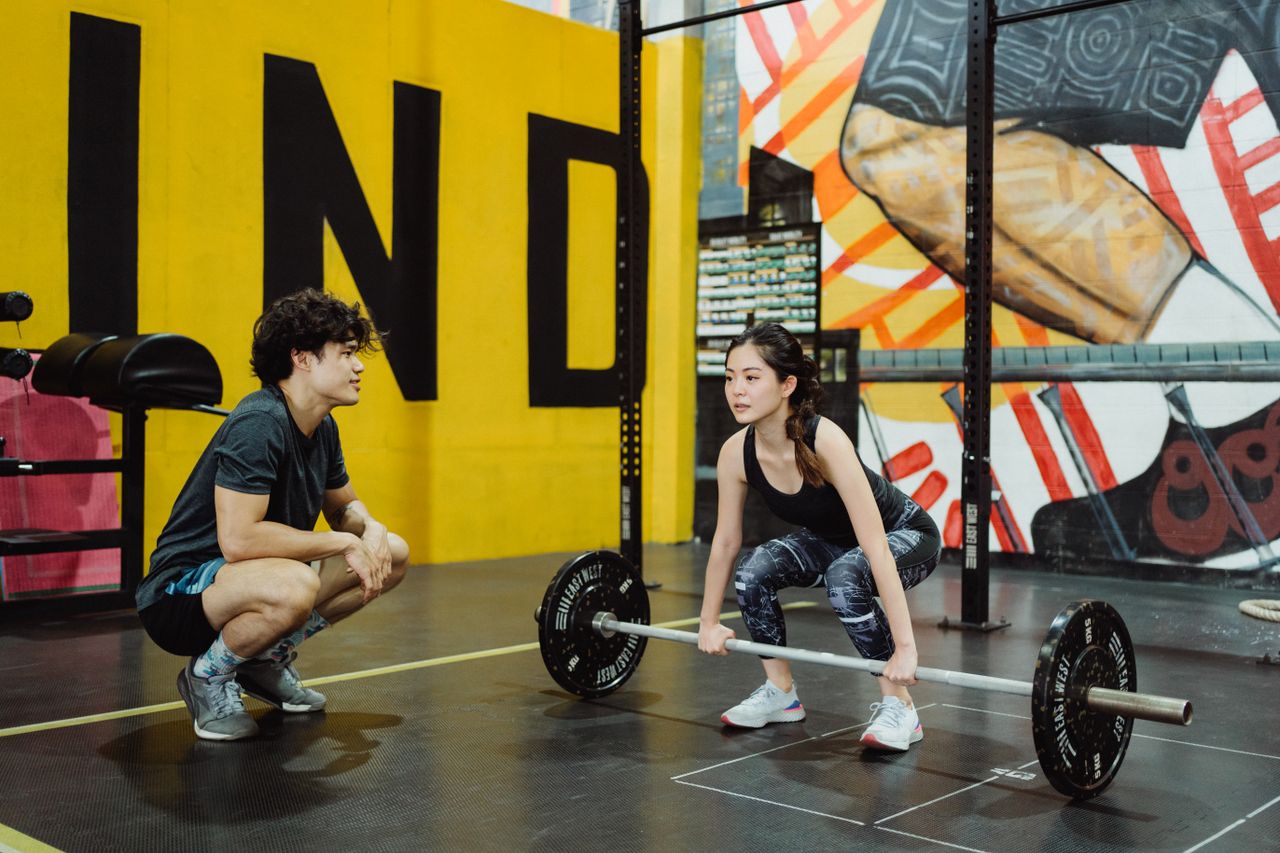 The Art of Drawing Readers In: Your attractive post title goes here A young woman lifting weights guided by a trainer in a colorful gym setting.